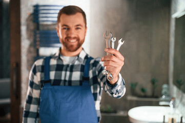 Holding wrenches. Plumber in blue uniform is at work in the bathroom