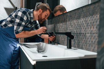 Side view. Fixing water tap. Plumber in blue uniform is at work in the bathroom