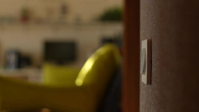 Close-up hands of unrecognizable young man turning light on and off by pushing switch on wall in dark bathroom, view of bright living room. Concept of home comfort. Shooting in slow motion.