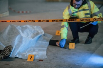 Knife is on the floor. Male detective is collecting evidence in a crime scene near dead body on the construction site