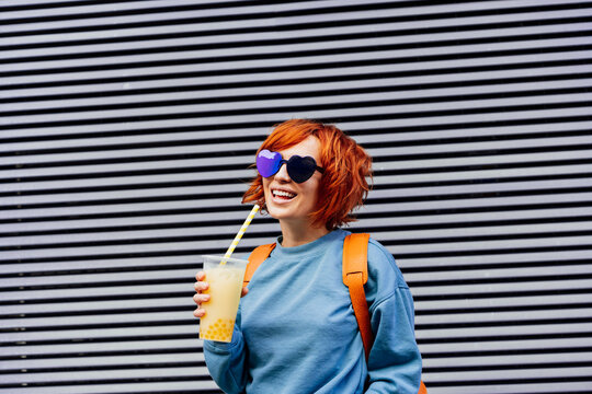 Smiling woman in heart shaped sunglasses and bright clothes drinking sugar flavored tapioca bubble tea near gray striped urban wall. Portrait of fashionable hipster girl. Street fashion. Copy space.