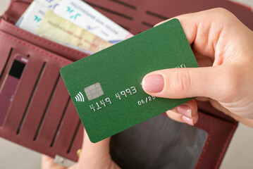 Woman's hands holding a green credit card and burgundy wallet with Ukrainian Hryvnias, close up....