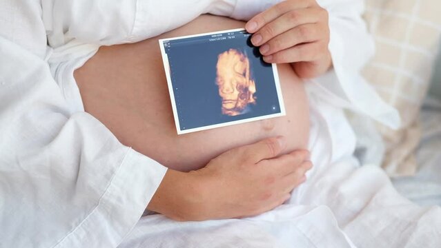 Unrecognizable pregnant woman wearing robe at home caressing,stroking belly, baby bump, holding ultrasound sonogram photo.Cropped, close-up shot.Happy,natural,calm,healthy pregnancy,relax,having rest.