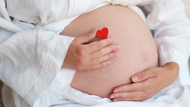 Unrecognizable pregnant woman wearing robe at home caressing,stroking belly, baby bump, holding red heart.Cropped, close-up shot.Happy pregnancy.Love and St Valentine day concept.