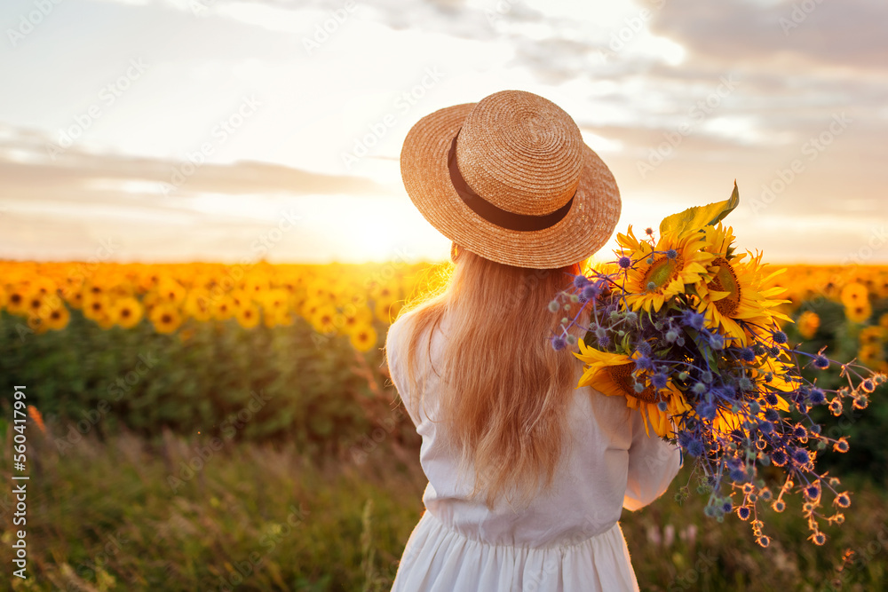 Wall mural Back view of woman enjoying view in blooming sunflower field at sunset with bouquet of flowers. Peace and freedom