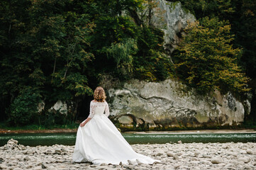 Romantic bride standing on the stony shore river in the mountains. The bride walking on nature. Back view. Country.