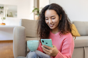 Attractive smiling Asian woman using smart phone while sitting on the sofa at home. Communication and video call concept.