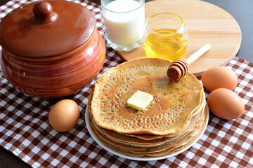 pile of russian blini on plate isolated with eggs, ceramic pot, honey and milk on plaid towel