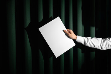 Hand in white shirt holding blank a format letter paper mockup template on a green fabric curtains...