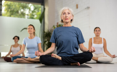 Peaceful mature female sitting in Lotus Pose Padmasana on mat during group yoga class in fitness studio