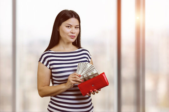 Young asian woman is taking money from the red wallet. Blurred windows with sunlight on the background.