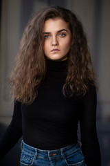 Naklejka premium Front photo of a feminity young woman with curly hair, wear black suitcase and jeans, looking at the side, posing near grey wall.