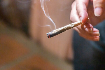 Finger holding lighted cannabis cigarette with smoke in dark room, with selective focus on cannibis...
