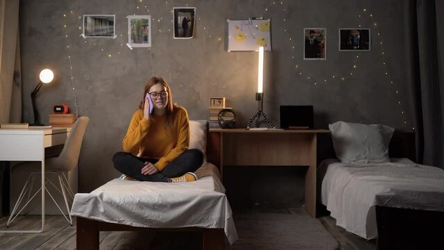 A student girl sits in a college dormitory on a bed in an orange sweater, talking on the phone using a smartphone.