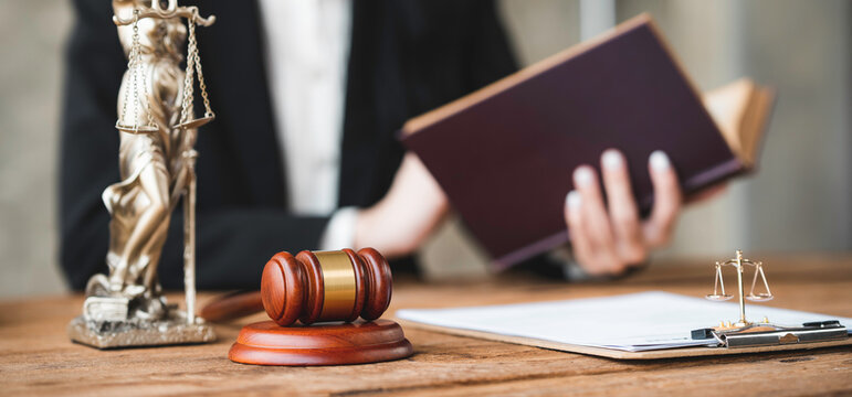 Asian Female Lawyer Or Legal Advisor Working With Scales Of Justice Sitting At Her Desk Holding A Book On The Scale Of Jurisprudence Concepts To Study And Find Information.