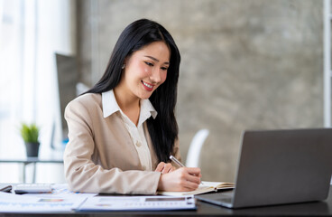 Confident Asian businesswoman sitting and taking notes in modern management concept with laptop computer in office happily.