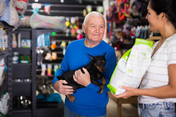 Old man choosing dog food for his miniature pinscher in pet shop. Saleswoman helping him with it.
