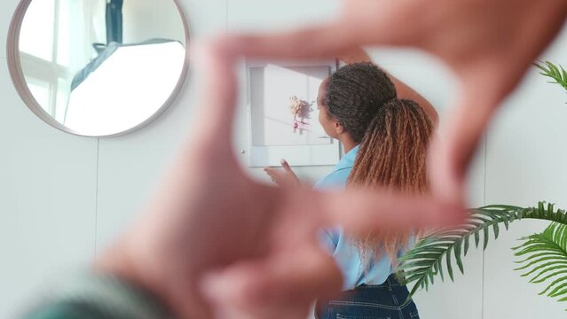 Young beautiful African American woman hangs picture on wall while guy uses fingers to determine best place for piece of art given by friends for housewarming located inside house with white walls