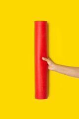 Mano de mujer sosteniendo una esterilla de yoga roja sobre un fondo amarillo brillante liso y aislado. Vista de frente y de cerca. Copy space. Formato vertical - obrazy, fototapety, plakaty