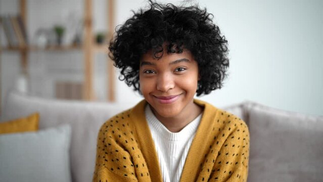 Beautiful portrait of african american girl with clean health skin and white toothy smile, sitting in sofa on home. Laughing, dreamy, pretty, young woman. Curly hair, style. Cozy interior and clothes.