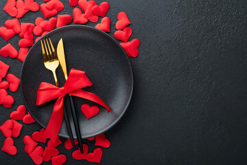 Valentines day romantic table setting. Empty or closeup of a dinner black plate, knife, fork and decorative silk hearts on black background. Holiday concept. Copy space for inscriptions.