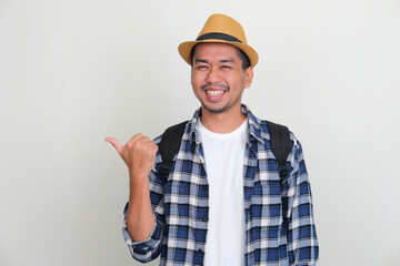 Asian backpacker smiling at the camera pointing to the right side using his thumb