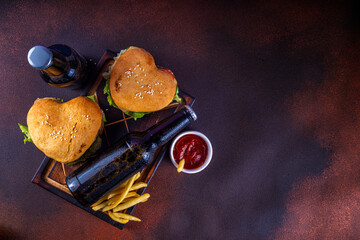 Valentine day Heart-shaped burgers. Two tasty cheeseburgers with french fries and beer bottles on dark table background. Idea for Valentine day dinner, dating, non-usual February 14 menu for couples