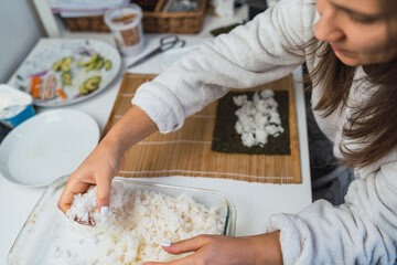 Woman taking a portion of rice from the pot to assemble a sheet of nori seaweed and prepare...