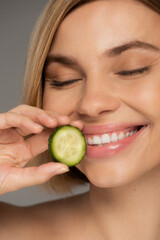 close up of cheerful woman with closed eyes holding sliced cucumber isolated on grey.
