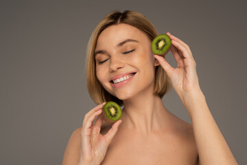 happy young woman with closed eyes holding kiwi halves isolated on grey.