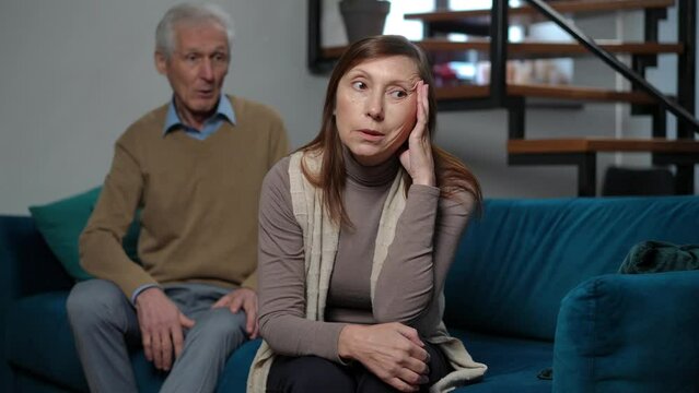 Rack focus from annoyed senior wife rolling eyes arguing with husband standing up leaving to irritated man sitting on couch in living room. Conflicts and relationship difficulties