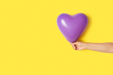 Obraz premium Mano de mujer sosteniendo un globo en forma de corazón de color morado sobre un fondo amarillo brillante liso y aislado. Vista de frente y de cerca. Copy space