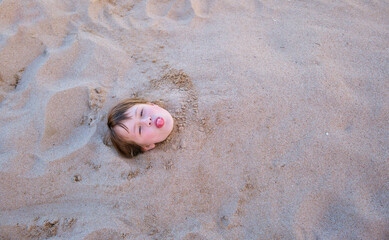 Young smiling child girl lying down covered with white sand on tropic beach on blue sky and ocean water background
