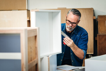 Fototapeta na wymiar Senior caucasian man specialist worker measuring white shelf furniture in wood workshop, Small business in wood furniture industry.