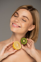 joyful woman with closed eyes holding kiwi fruit and lemon isolated on grey.