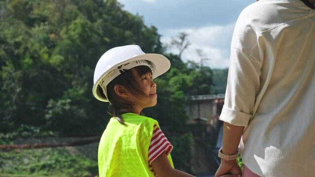 Engineer mother holding her daughter's hand and smiling at each other against the background of a dam with a hydroelectric power station. Concepts of renewable energy and love of nature and family.