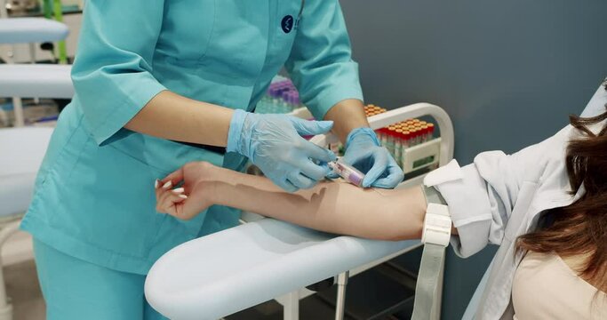 From above handheld shot of female assistant in uniform inserting syringe needle into vein and putting alcohol wipe on arm while taking blood sample of woman during work in clinic.