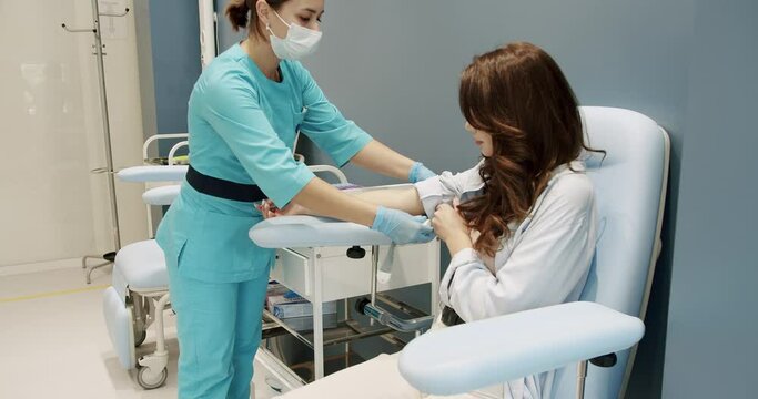 Crop nurse taking blood sample of patient. female assistant in uniform inserting syringe needle into vein and putting alcohol wipe on arm while taking blood sample of woman during work in clinic.