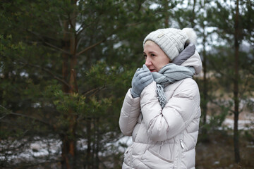 Portrait of beautiful happy senior retired elderly woman is freezing walking outdoors in winter snowy park or forest, wood, at cold frosty day in hat, scarf, gloves, in snow, trembles and smoke 