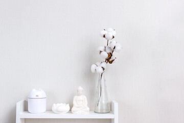 Fototapeta na wymiar Buddha figurine, air humidifier, lotus candlestick and vase with branch of cotton on white shelf. Concept of comfortable living in the house. Selective focus, copy space