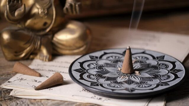 Burning incense cone with smoke on a soapstone holder with mandala, Ganesha statue in the background