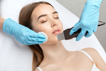 The doctor-cosmetologist makes the ultrasound cleaning procedure of the facial skin of a beautiful, young woman in a beauty salon. Cosmetology and professional skin care.	
