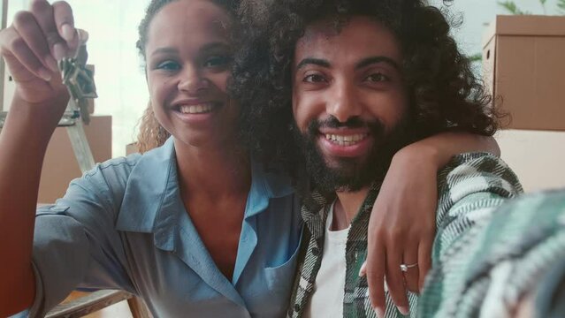 Young inspired multiracial couple Arabian man and African American woman showing off keys to buying house take joint selfie for family photo album or social networks hugging posing in room with boxes