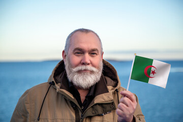 Man holding Algeria flag. Portrait of older man with a national Algerian flag. Visit Algeria. Older man 50 55 60 years old with gray beard outdoors travelling in winter. Travel to Algeria. - 560508537