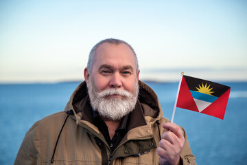 Man holding Antigua and Barbuda flag. Portrait of older man with a national flag. Visit Antigua and Barbuda. Older man 50 55 60 years old with gray beard outdoors travelling in winter. - 560508550