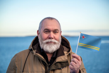 Man holding Aruba flag. Portrait of older man with a national Aruba flag. Visit Aruba concept. Older man 50 55 60 years old with gray beard outdoors travelling in winter. - 560508571