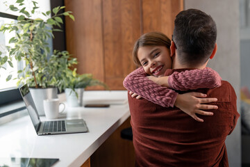 Caring father and his loving daughter embracing in a home office during his pause from work on a laptop. Little girl is smiling and looking at the camera. Childhood and fatherhood moments.