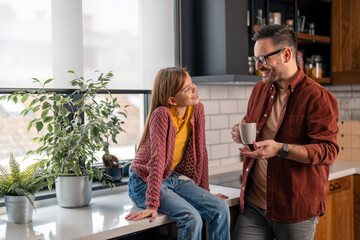 Dad spending time with his daughter at home, drinking a coffee, having a conversation with her, gesturing with hand. Daughter is sitting on a kitchen counter and smiling at him.