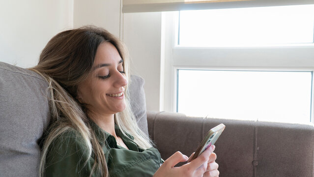 flirty woman smiling to a message on the phone. Young adult blonde woman typing on phone. Beautiful charming girl texting on smart phone at home.