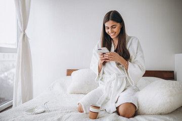 Young woman in bed in bathrobe with phone drinking coffee - obrazy, fototapety, plakaty